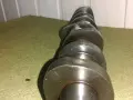 Metalowy, wytarty wałek rozrządu leży na zielonym materiale. Widać ślady zużycia na jego krzywkach.