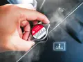Dłoń dotyka gorącego korka chłodnicy, z którego unosi się para. Widoczny symbol ostrzegawczy i napis "DANGER WARNING". Kuhlmittel stand jest wysoki.