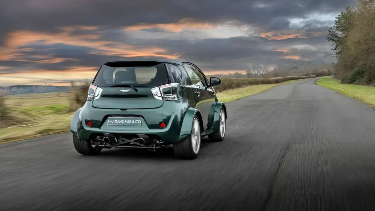 Zielony Aston Martin Cygnet V8 z szerokimi błotnikami pędzi po wiejskiej drodze pod dramatycznym, zachodzącym słońcem.
