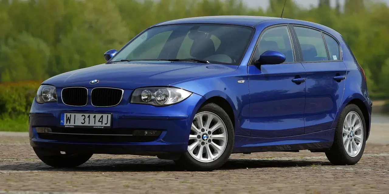 Błękitne BMW E87 z polskimi tablicami WI 3114J, z nowymi żarówkami w reflektorach, zaparkowane na bruku.