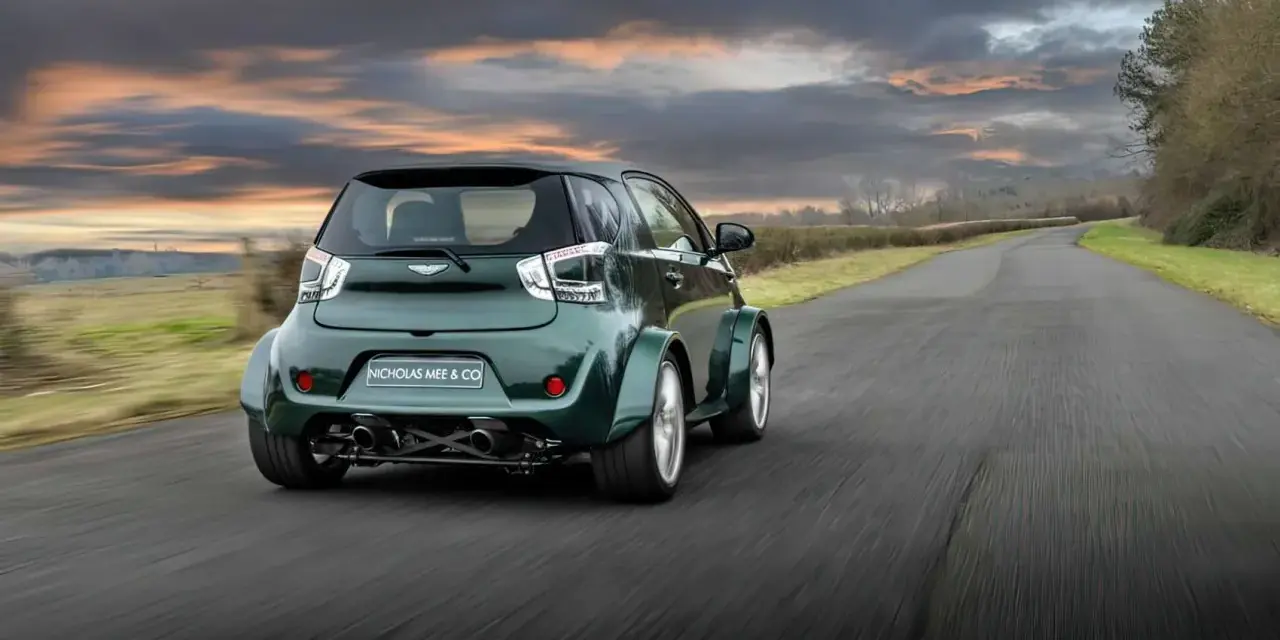 Zielony Aston Martin Cygnet V8 z szerokimi błotnikami pędzi po wiejskiej drodze pod dramatycznym, zachodzącym słońcem.