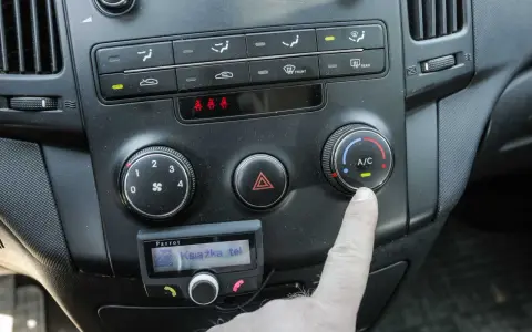 Palec naciska przycisk A/C w samochodzie, włączając klimatyzację. Widoczne pokrętła nawiewów i panel sterowania.