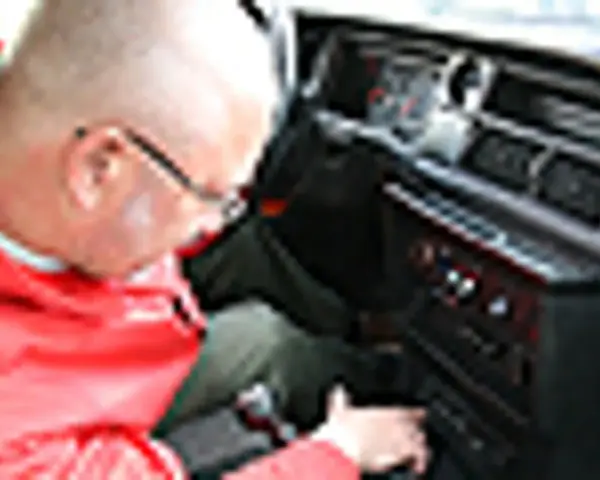 Mężczyzna w czerwonej kurtce pochyla się nad deską rozdzielczą, szukając rozwiązania, **co zrobić żeby radio nie szumiało**.