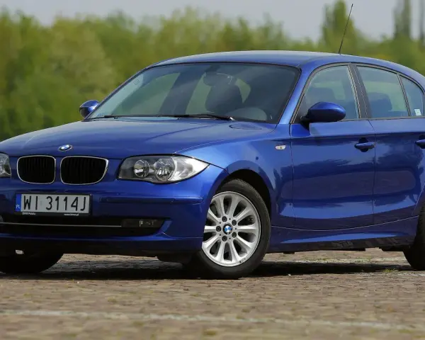 Błękitne BMW E87 z polskimi tablicami WI 3114J, z nowymi żarówkami w reflektorach, zaparkowane na bruku.