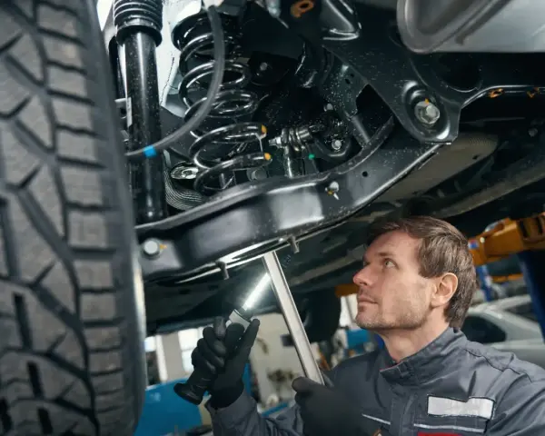 Mechanik bada elementy zawieszenia samochodu, oświetlając je latarką. Widoczna sprężyna i amortyzator.