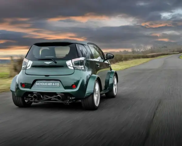 Zielony Aston Martin Cygnet V8 z szerokimi błotnikami pędzi po wiejskiej drodze pod dramatycznym, zachodzącym słońcem.