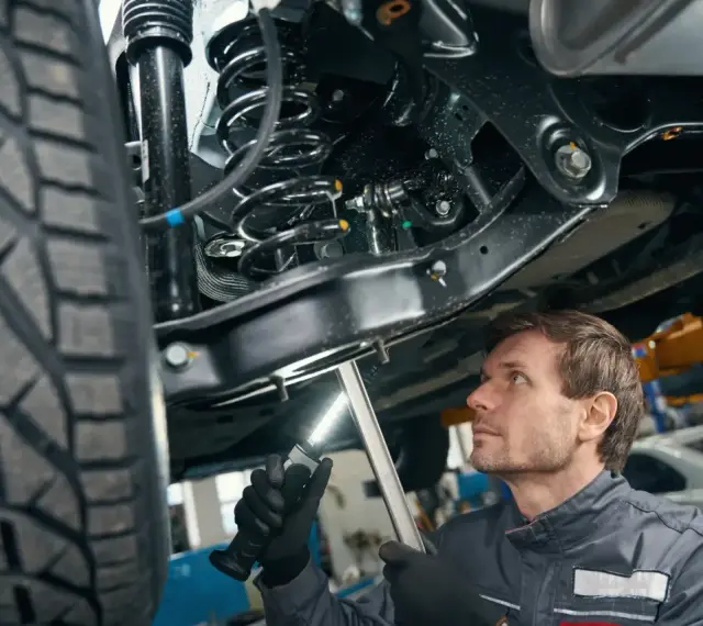 Mechanik bada elementy zawieszenia samochodu, oświetlając je latarką. Widoczna sprężyna i amortyzator.