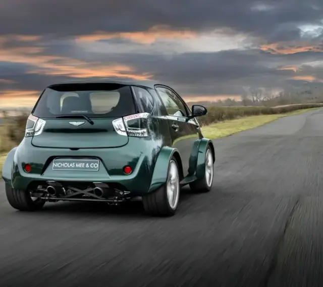 Zielony Aston Martin Cygnet V8 z szerokimi błotnikami pędzi po wiejskiej drodze pod dramatycznym, zachodzącym słońcem.