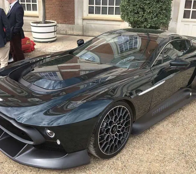 Elegancki, czarny Aston Martin Victor błyszczy na tle ceglanego budynku. Jego aerodynamiczna linia i unikalne felgi przyciągają wzrok.