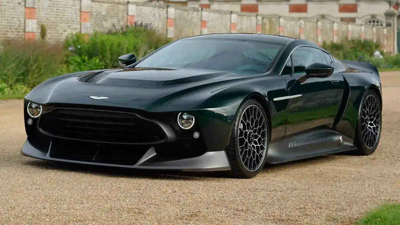 Elegancki, ciemnozielony Aston Martin Victor z unikalnymi felgami, zaparkowany na żwirowej drodze przed zabytkowym budynkiem.