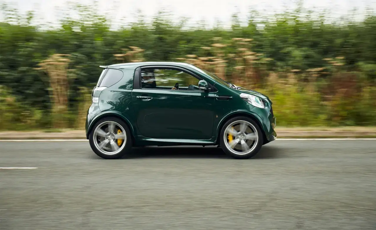 Zielony Aston Martin Cygnet V8 pędzi po drodze, otoczony rozmazaną zielenią.