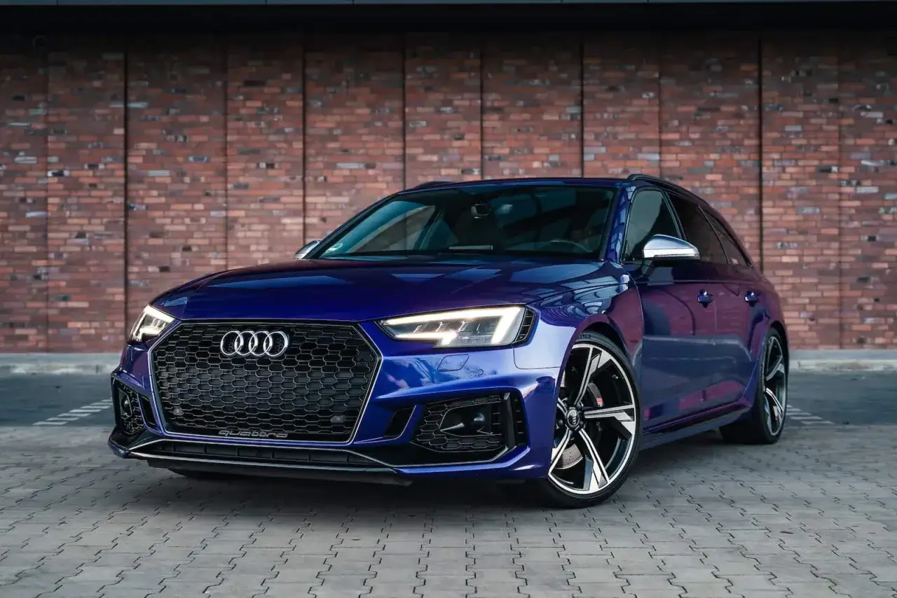 Błękitne Audi RS4 Avant z czarnym grillem i felgami stoi przed ceglaną ścianą.
