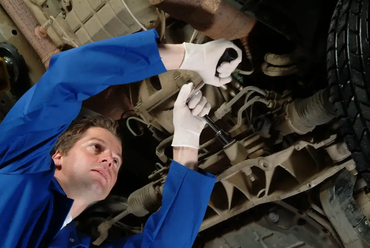 Mechanik w niebieskim kombinezonie i białych rękawiczkach dokonuje wymiany przegubu w samochodzie.
