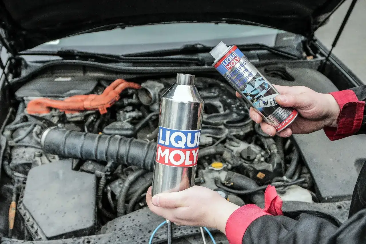 Dłonie trzymają preparaty Liqui Moly do czyszczenia katalizatora. Silnik samochodu w tle.