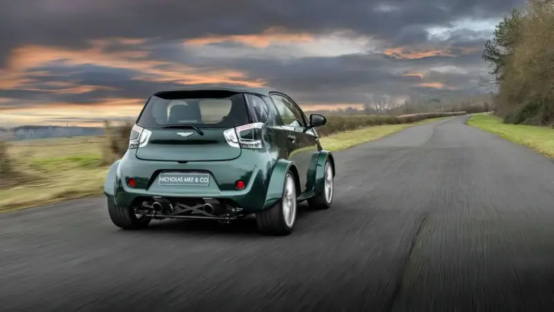 Zielony Aston Martin Cygnet V8 z szerokimi błotnikami pędzi po wiejskiej drodze pod dramatycznym, zachodzącym słońcem.