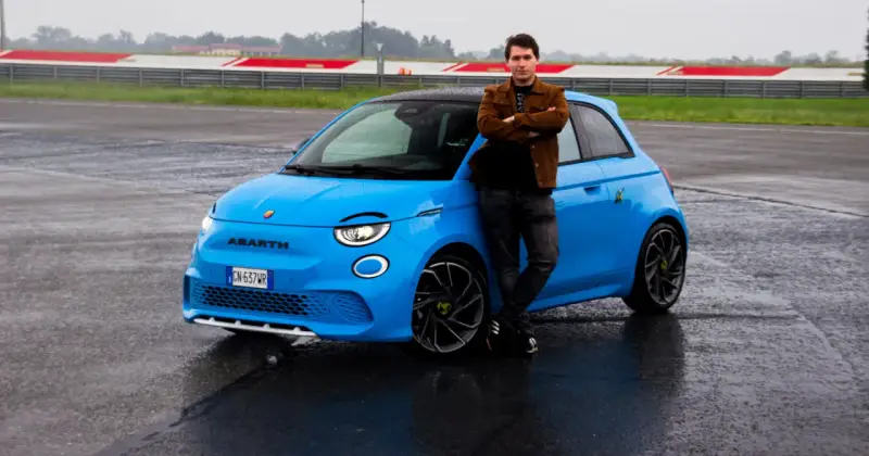 Mężczyzna opiera się o jaskrawo niebieski Abarth 500e na mokrym torze wyścigowym.